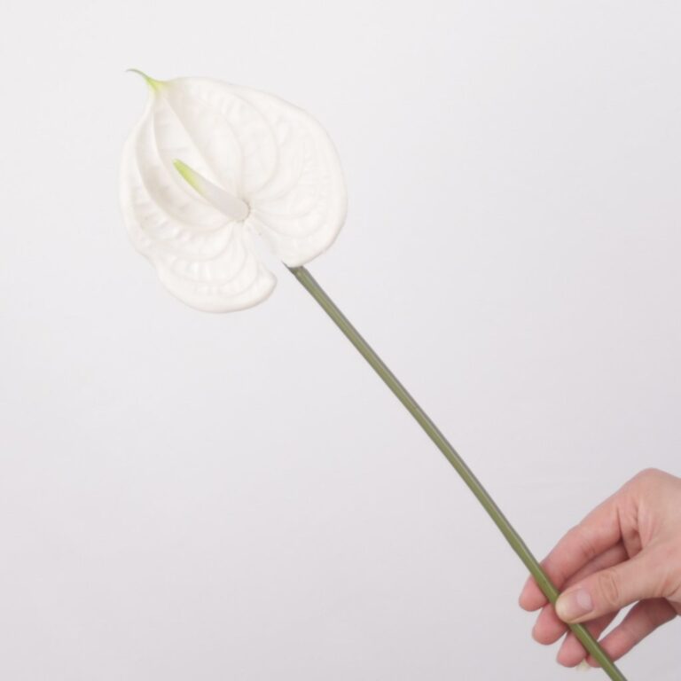 Close-up of a single artificial white anthurium flower with a realistic waxy texture and a yellow-green spadix, on a green stem against a light gray background.