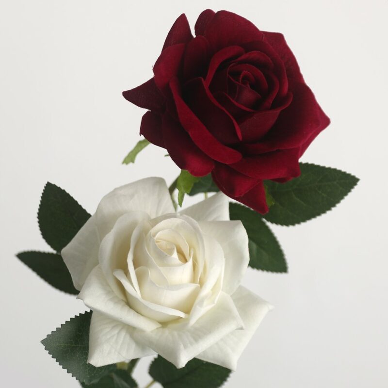 A pair of realistic artificial roses: one deep dark red (maroon) blooming rose head positioned above one creamy white blooming rose head, both with realistic green leaves on a stem, against a plain gray background.