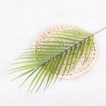Realistic faux tropical green leaves arranged on a woven beige decorative coaster or mat.