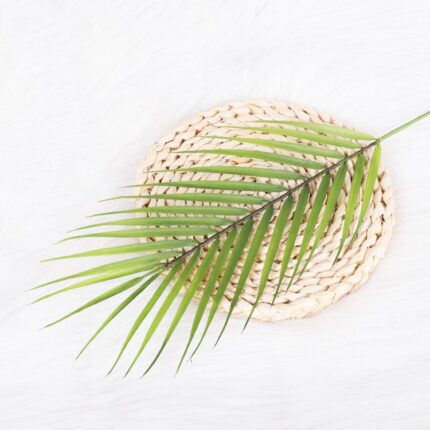 Realistic faux tropical green leaves arranged on a woven beige decorative coaster or mat.