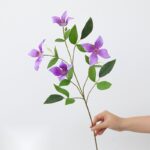 Silk clematis flower branch featuring four vibrant purple blooms and lush green foliage.