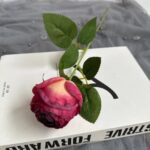Realistic artificial silk rose, featuring deep burgundy to blush pink hues, resting on a book cover with leaves.