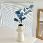 Artificial dusty blue foliage arrangement in a ceramic vase displayed on a white tabletop.