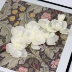 White silk artificial orchid arrangement of 8 heads displayed on a patterned floral textile backdrop.