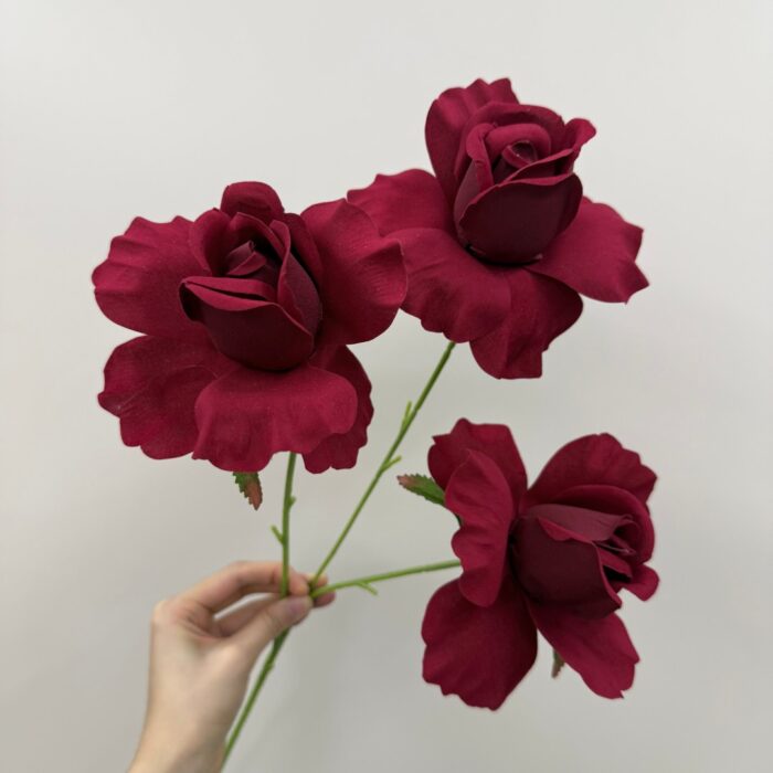 A hand holding a stem with three realistic burgundy artificial roses against a clean background.